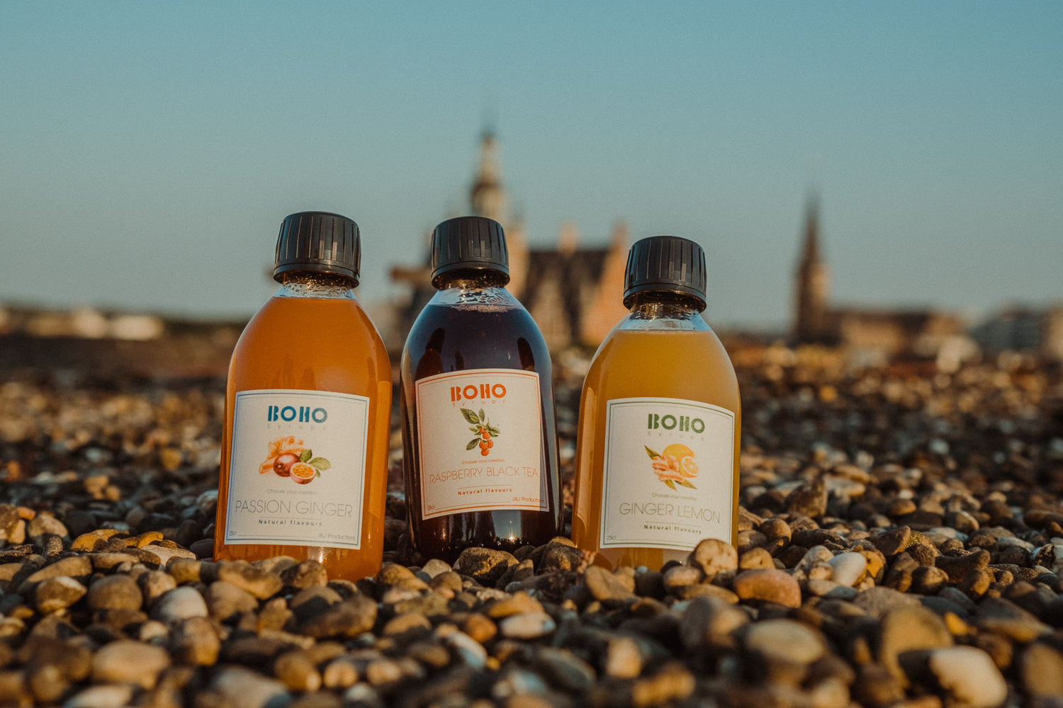 Drie flesjes BOHO fruitsiroop rechtop in kiezels met een blauwe lucht op de achtergrond