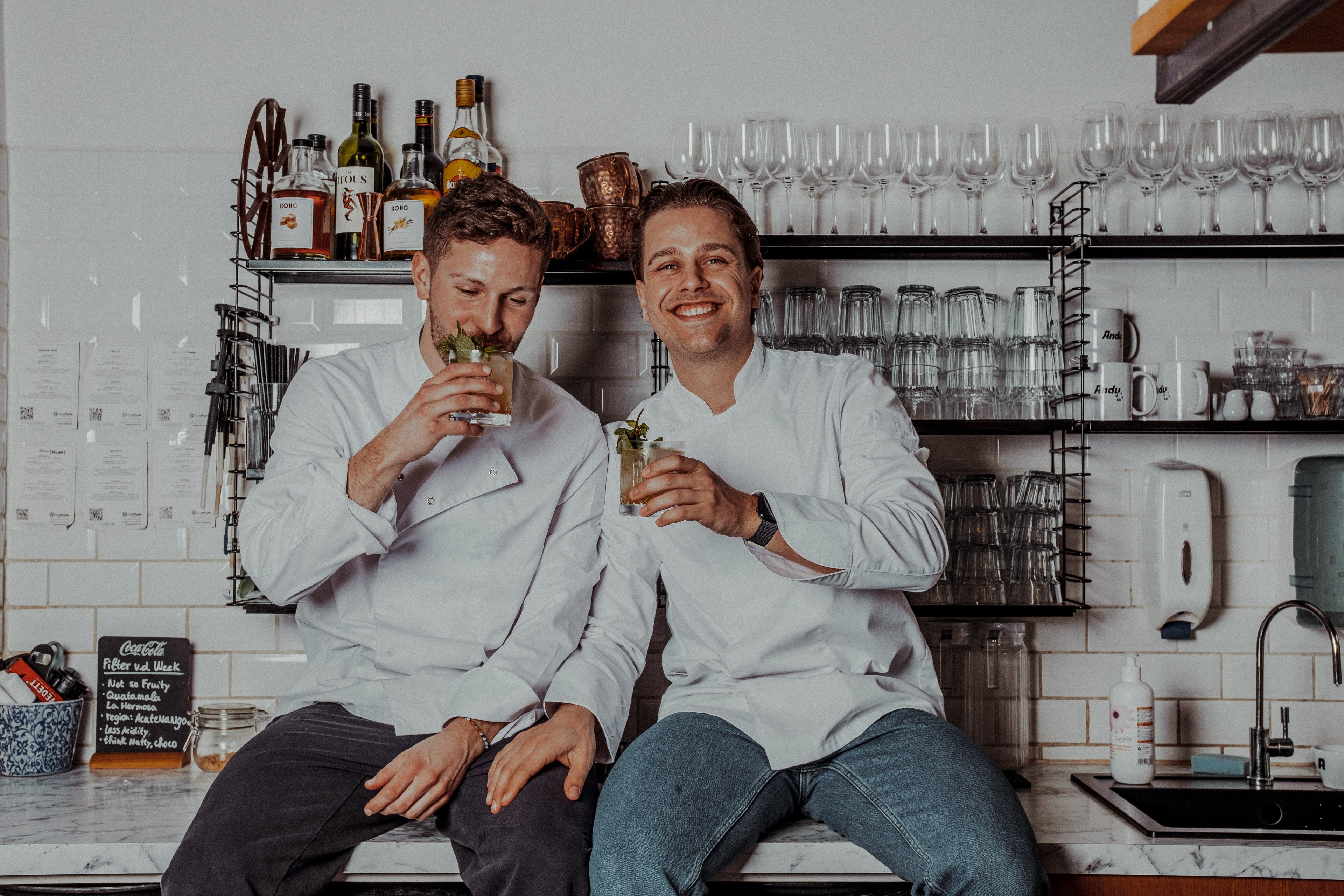 Joeri en Jef van BOHO Syrups zitten lachend op de toonbank in hun productiejas en klinken samen met een drankje – ambachtelijk en gezellig merkverhaal