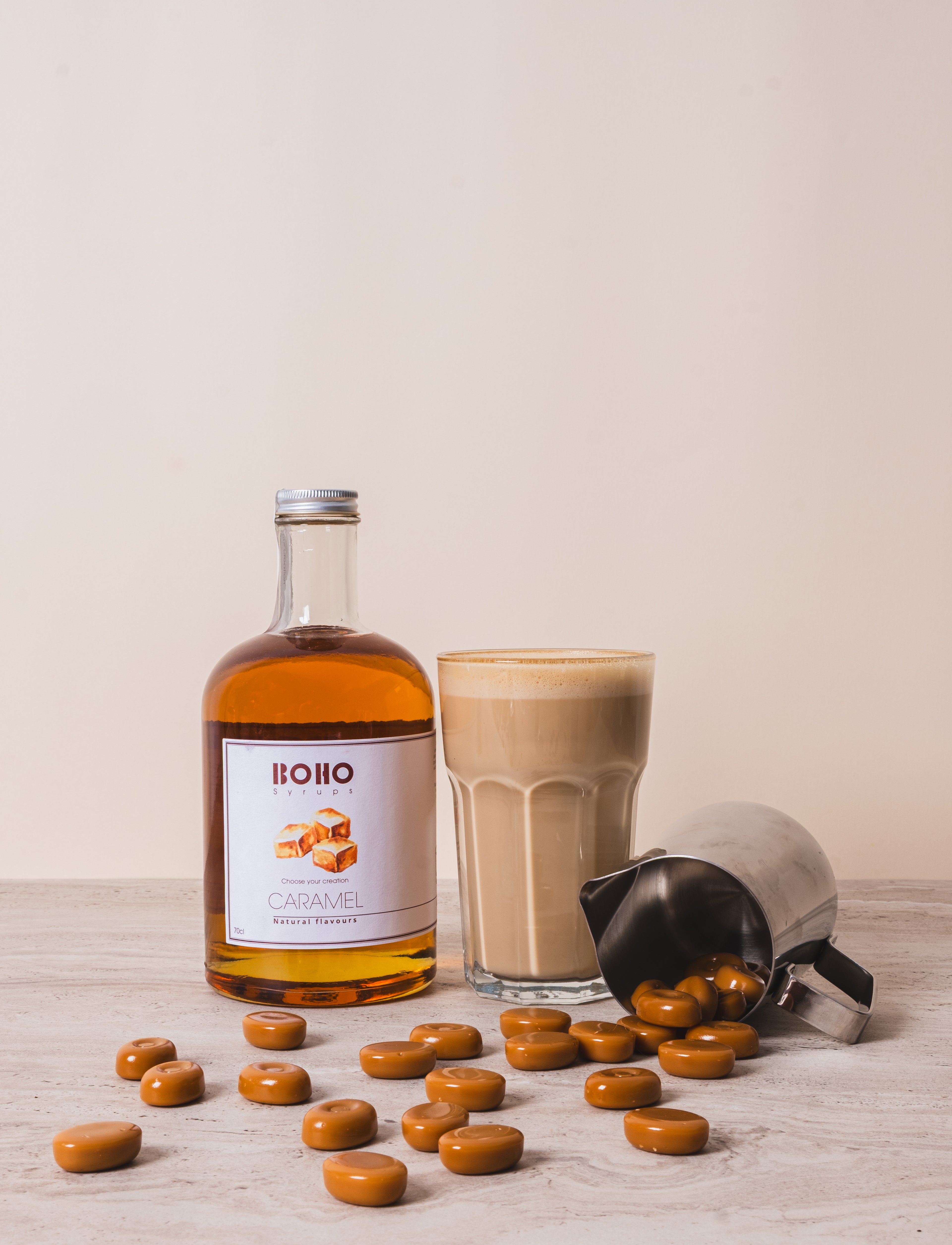 Fles BOHO karamel siroop met een latte macchiato ernaast en karamel snoepjes op de voorgrond – zoete en romige koffiesfeer