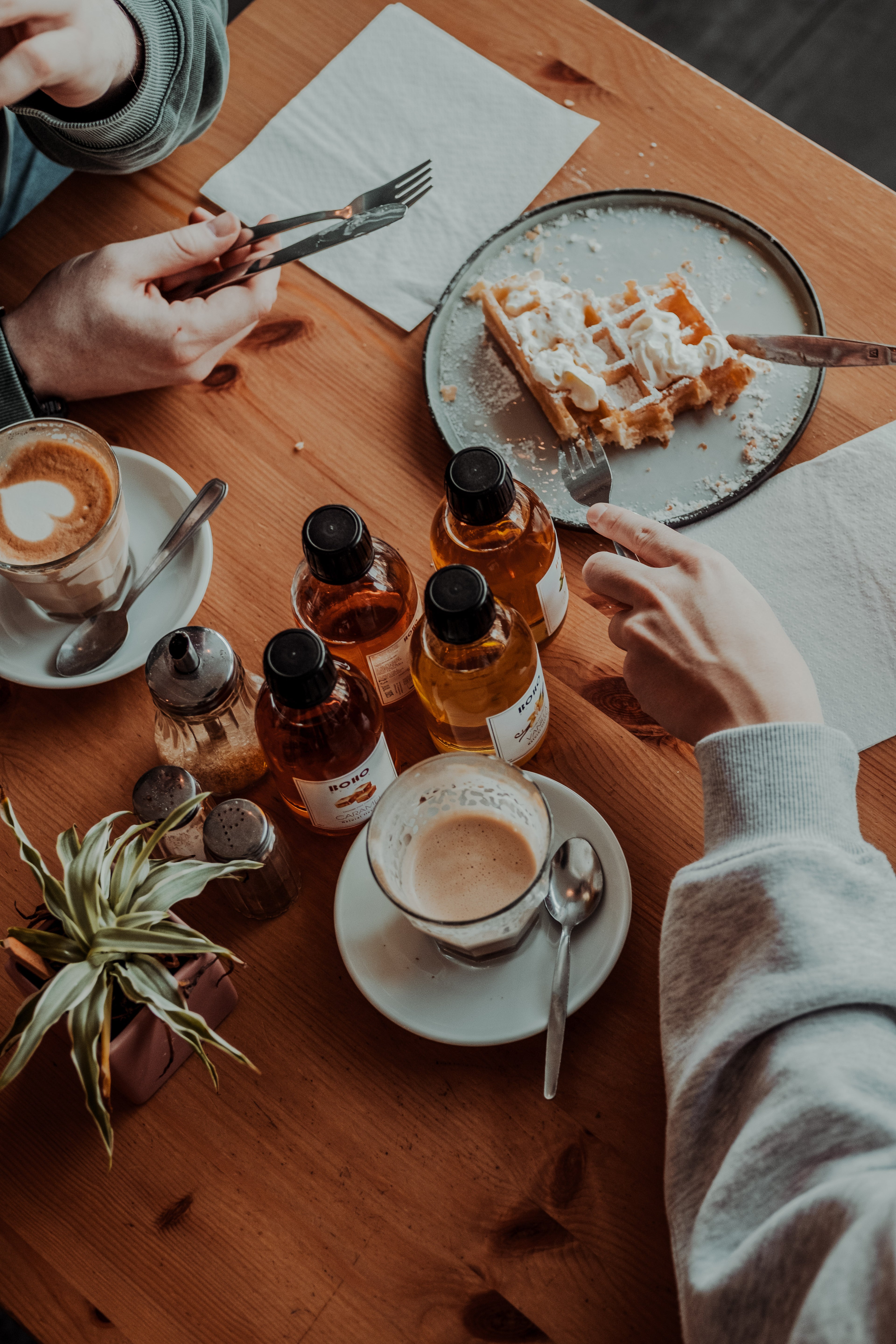 Vier flesjes BOHO koffiesiroop met cappuccino’s en een half opgegeten wafel op een houten tafel.